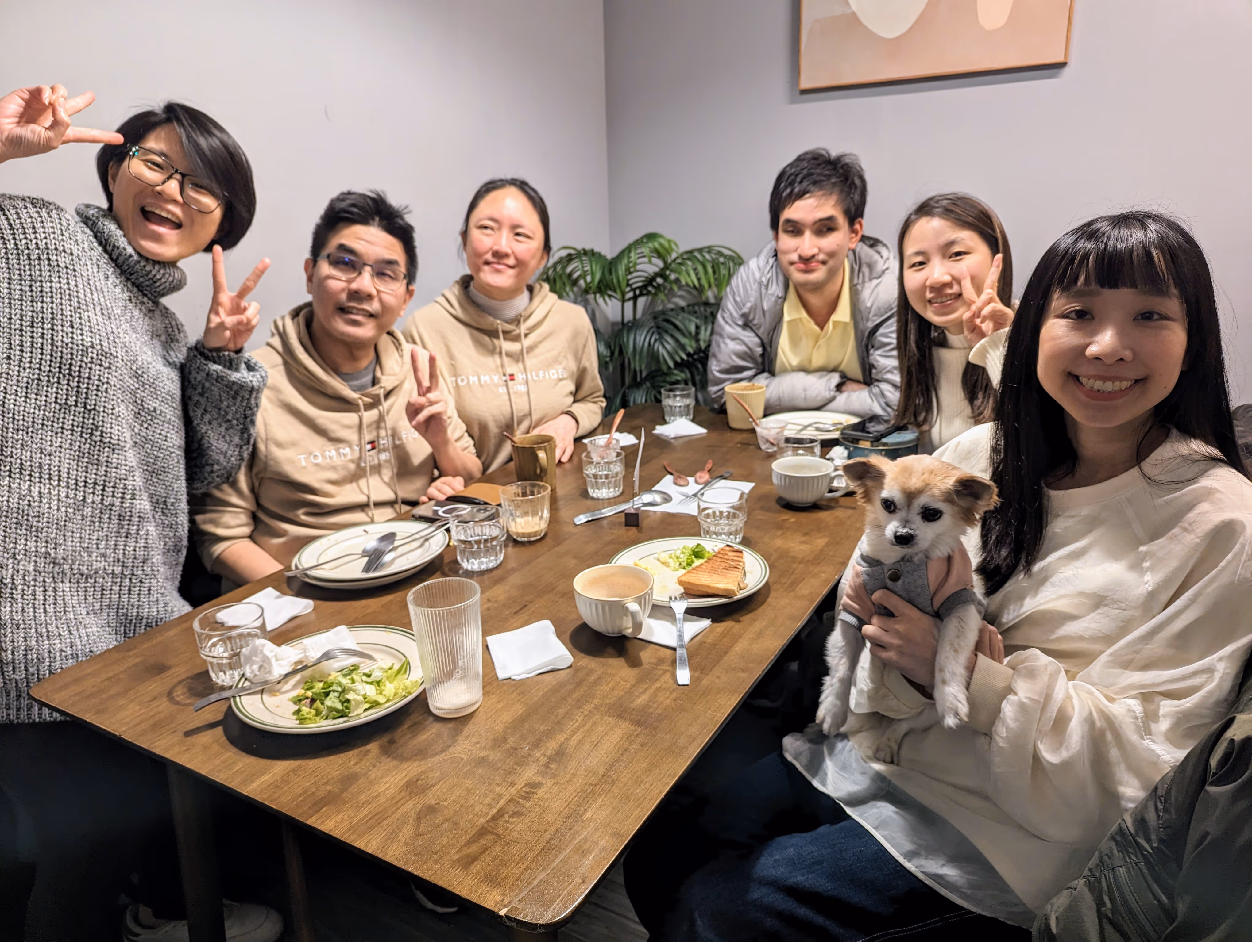 這張照片中有六個人在一張餐桌旁合影。左邊站著一位穿著灰色毛衣的女士，她戴著眼鏡，微笑著比出勝利手勢。坐在她旁邊的是一位穿著棕色連帽衫的男士，他也比出勝利手勢，旁邊坐著一位穿著相同連帽衫的女士。桌子的另一邊坐著三個人，其中一位女士抱著一隻小狗，微笑著看向鏡頭。桌上有一些餐具、飲料和食物，背景中有一幅掛畫和一些綠色植物。整體氛圍輕鬆愉快。