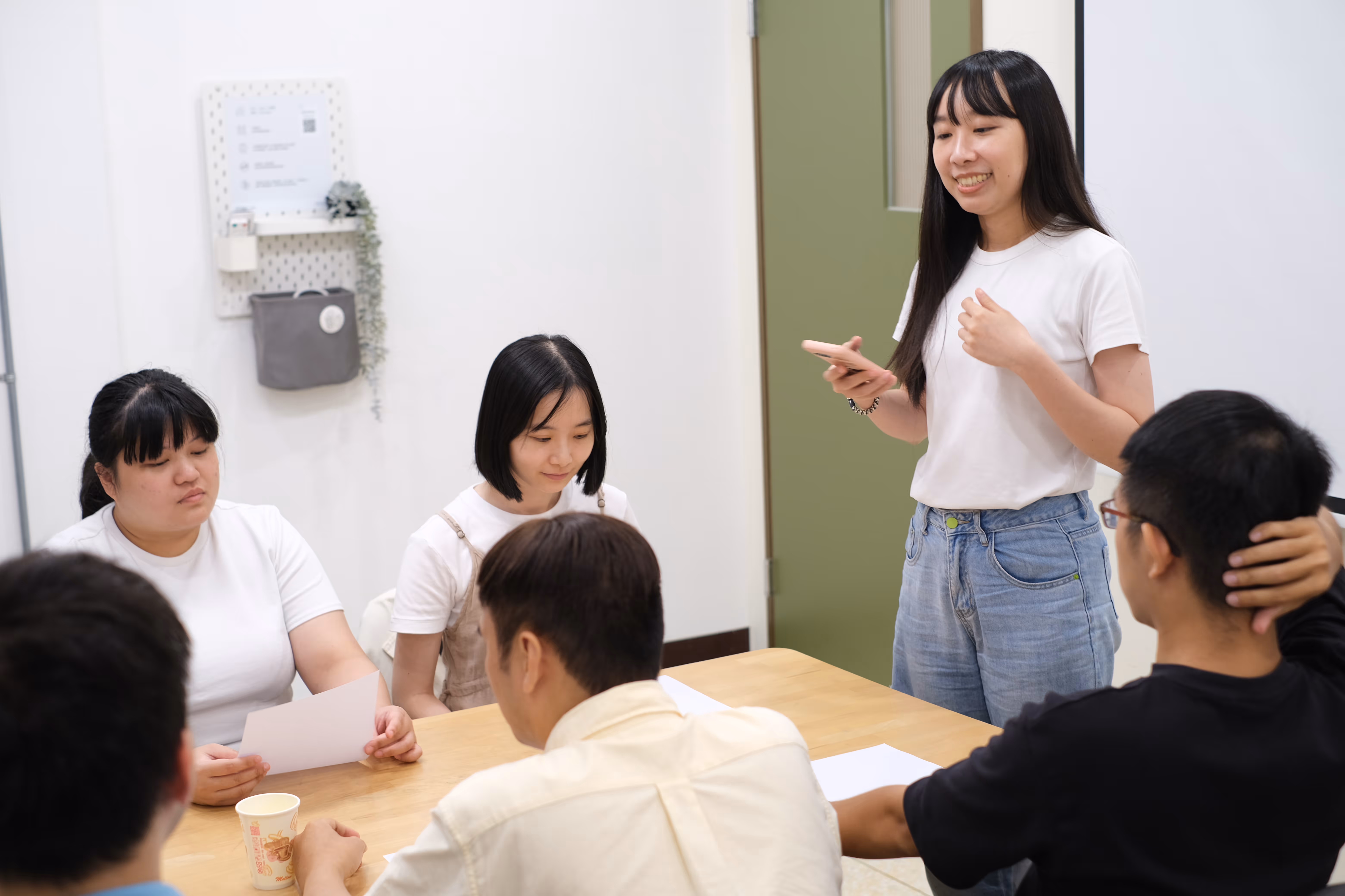 這張照片顯示一個會議或小組討論的場景。六個人圍坐在一張木桌旁，其中一位女性站著，似乎正在發言或做報告。她穿著白色上衣和牛仔褲，手裡拿著手機，面帶微笑。坐著的其他人中，有兩位女性和三位男性。左邊的女性穿著白色上衣，手中拿著一張紙，似乎在聆聽。另一位女性穿著白色上衣和吊帶裙，低頭看著桌子。桌上有一些紙張和一個紙杯。背景中有一扇綠色的門和一個掛在牆上的小架子，上面有一些裝飾物。整體氛圍看起來輕鬆而專注。