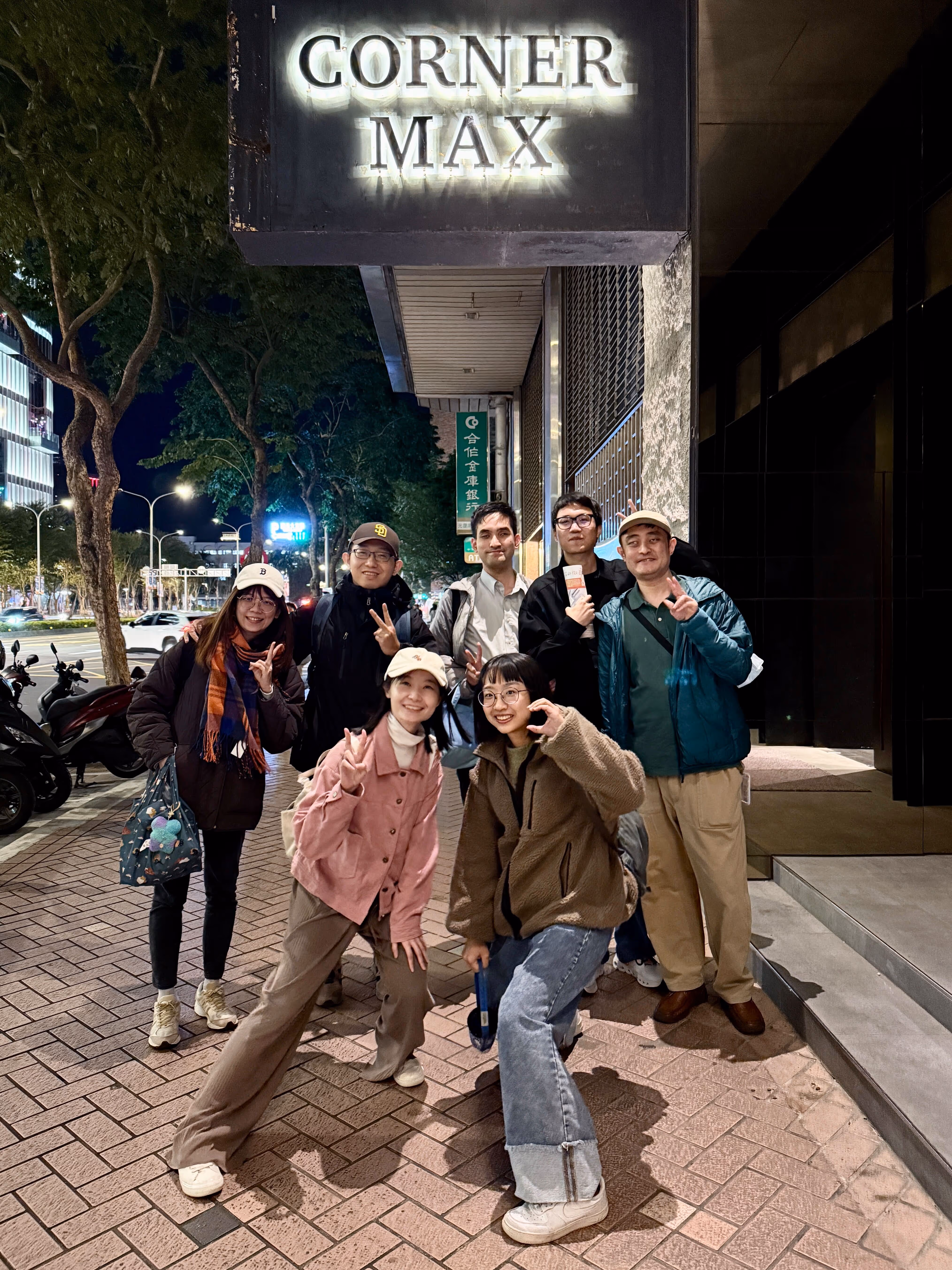 這張照片顯示了一群人在夜晚於街道上合影。背景中有一個招牌，上面寫著「CORNER MAX」。共有七個人，他們大多穿著休閒服裝，並且看起來很開心。前排有三位女性，兩位穿著帽子，並且做出比V的手勢。後排有四位男性，其中一位拿著飲料，大家都面帶微笑。背景中可以看到一些樹木和停放的摩托車，還有街道的燈光。整體氛圍輕鬆愉快。