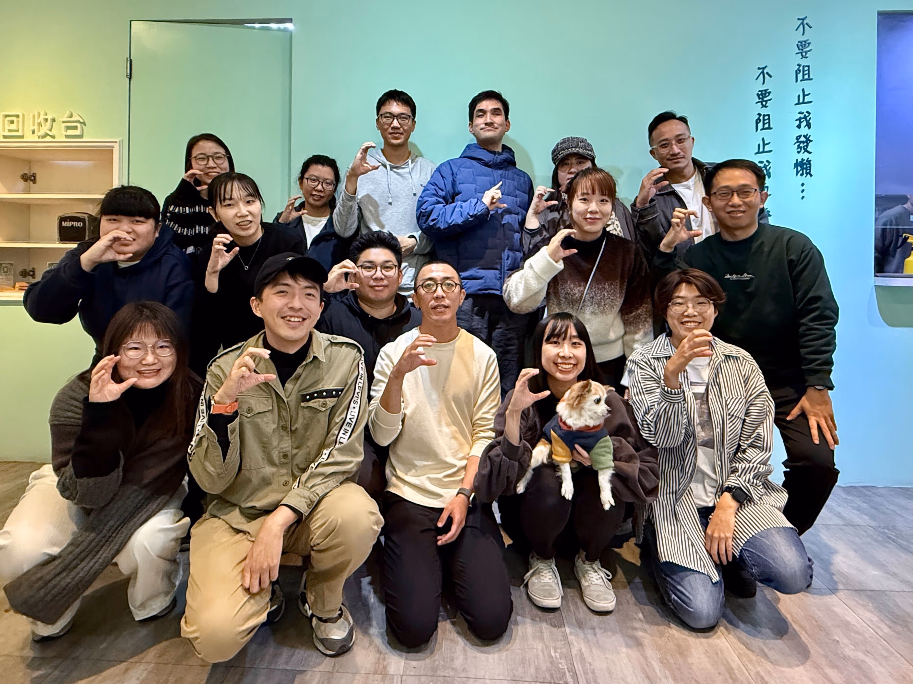 這是一張團體合照，照片中有17個人和一隻小狗。大家分成兩排站立和蹲坐在一個淺綠色的背景前。每個人都在微笑，並用手比出一個"C"的手勢。背景牆上有兩行垂直的文字，寫著「不要阻止我發懶... 不要阻止我發懶...」。左邊有一個小櫃子，上面寫著「回收台」。整體氣氛輕鬆愉快。